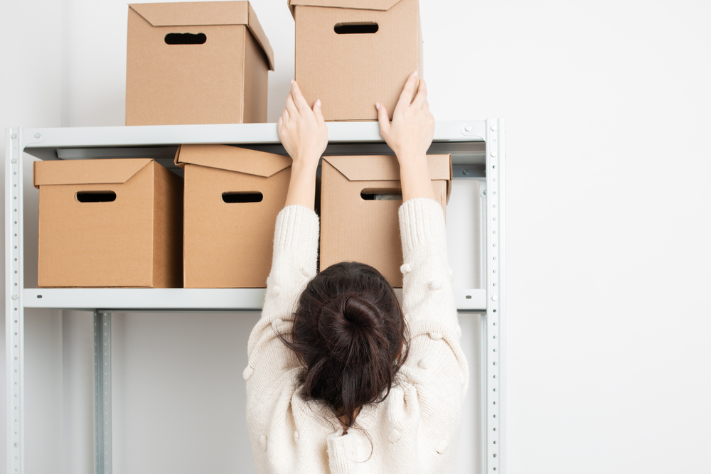 Woman reaching for a box on the top row of a shelf.