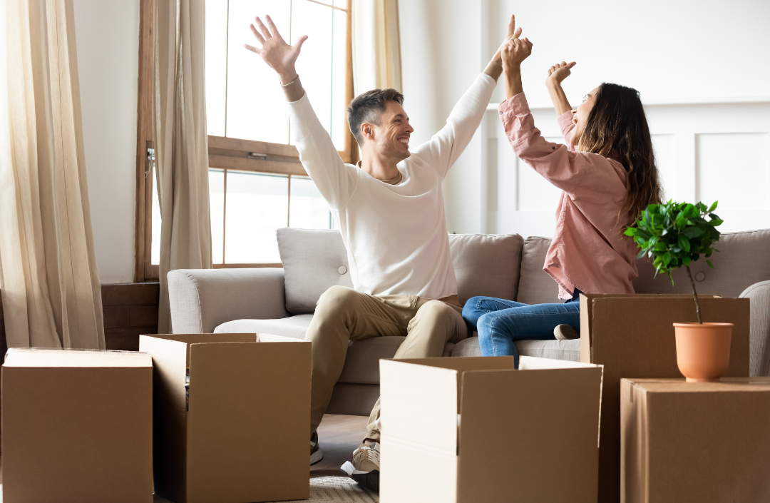 Couple sitting on a couch celebrating a successful move with cardboard boxes sitting on the floor in front of them.