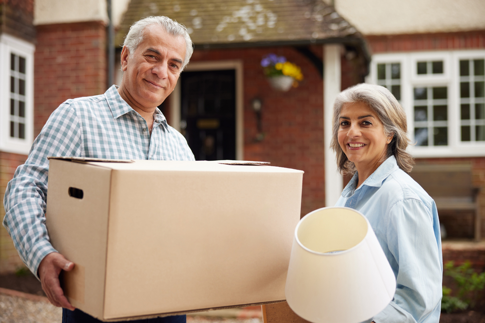 A couple standing outside of a house holding a box and a lamp.