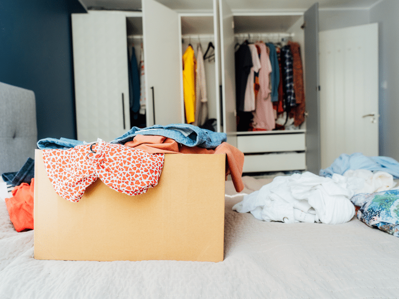 A room with clothes on the bed and a box of clothes.