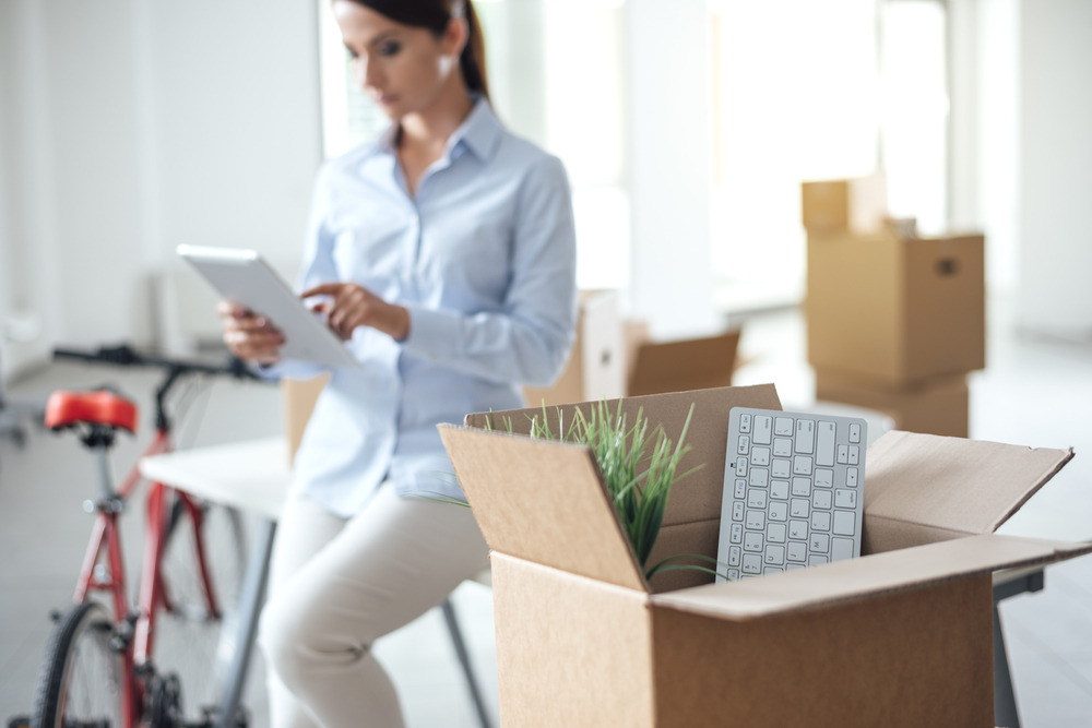 A woman sitting on a char looking at an ipad with office equipment in boxes.