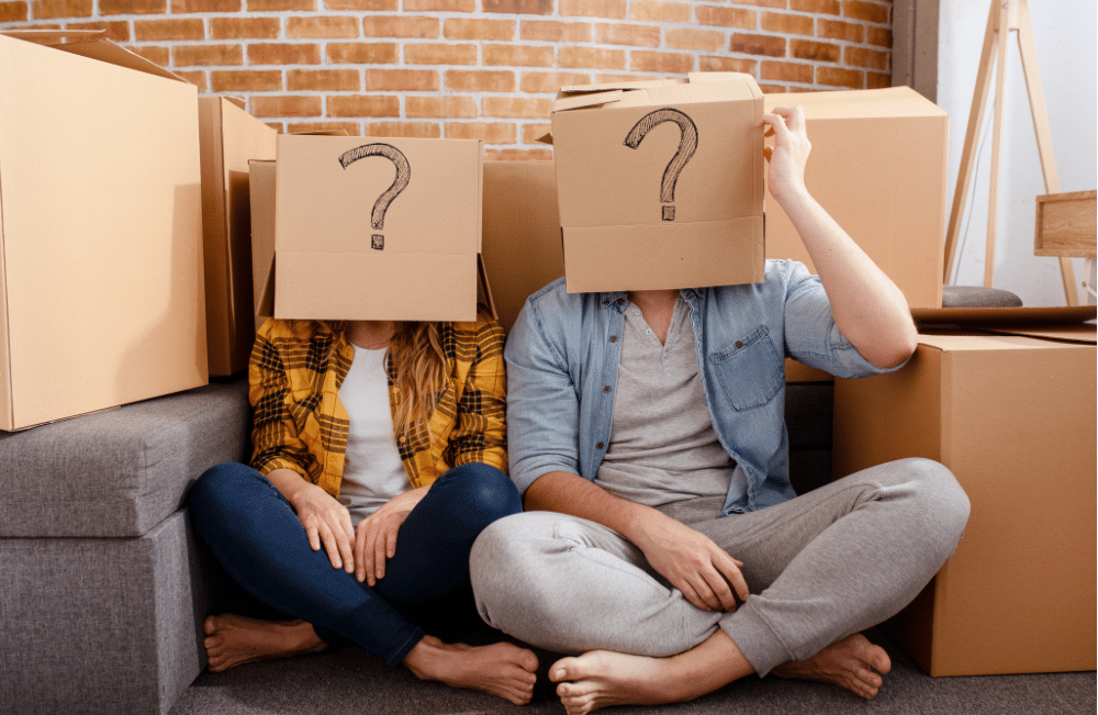 Man and woman sitting on the floor with cardboard boxes on their heads with question marks drawn on them.