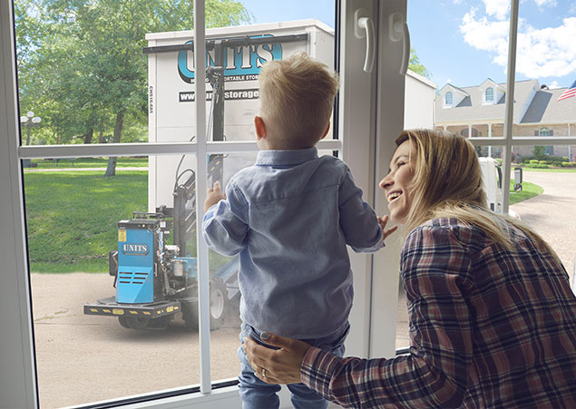 Woman smiling at her young son who is looking out the window at a UNITS Moving and Portable Storage ROBO-UNIT moving a storage container into their driveway.
