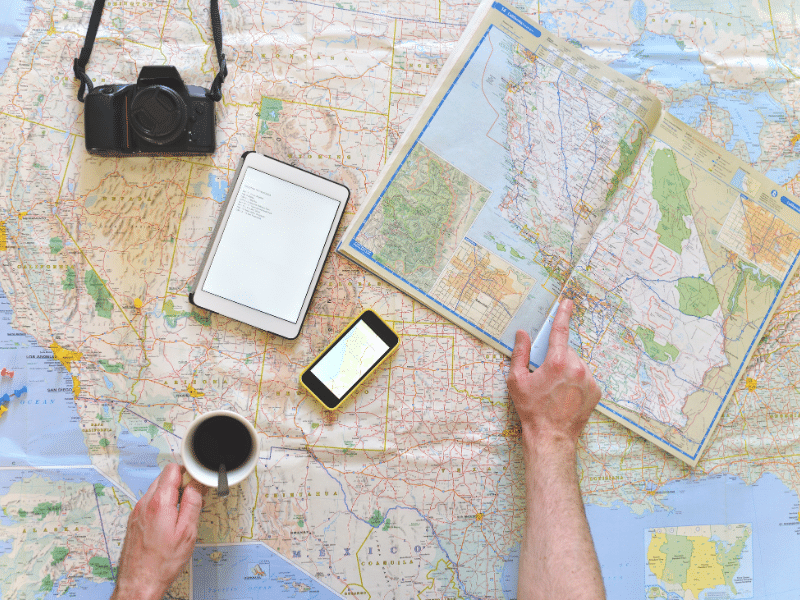Person going over a map of the United States with a phone, tablet, camera and a cup of coffee sitting on the map.