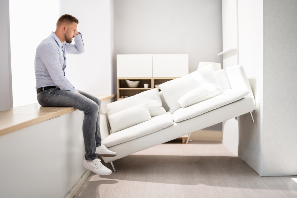 A man sitting on a ledge with his hand on his head having trouble moving a couch.