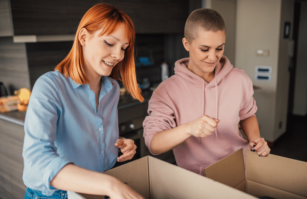 Two women standing in a kitchen packing items into boxes.