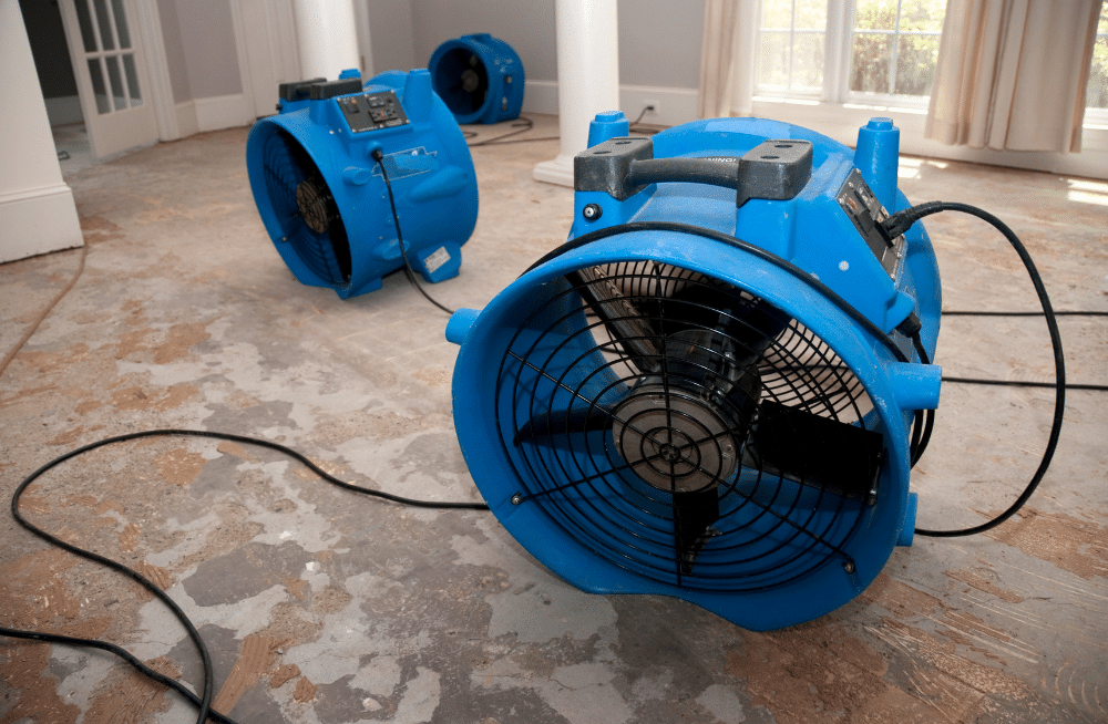 Three large fans blowing in a room with unfinished floors.