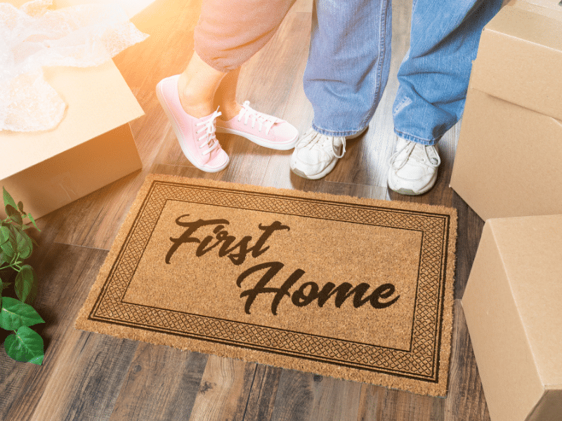 A man and a woman standing in front of a doormat that reads First Home.