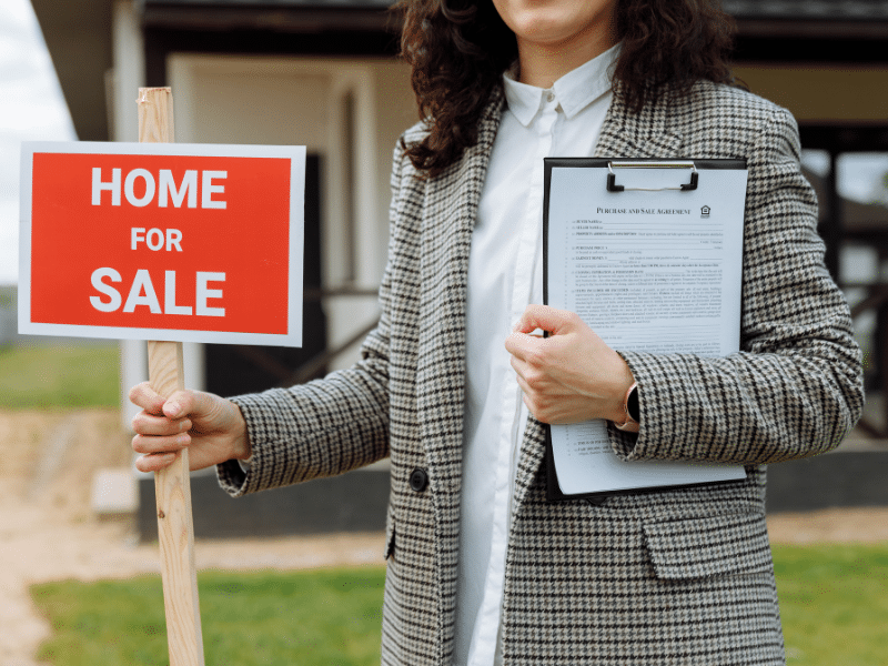A woman holding a Home For Sale sign in one hand and a contract in the other.