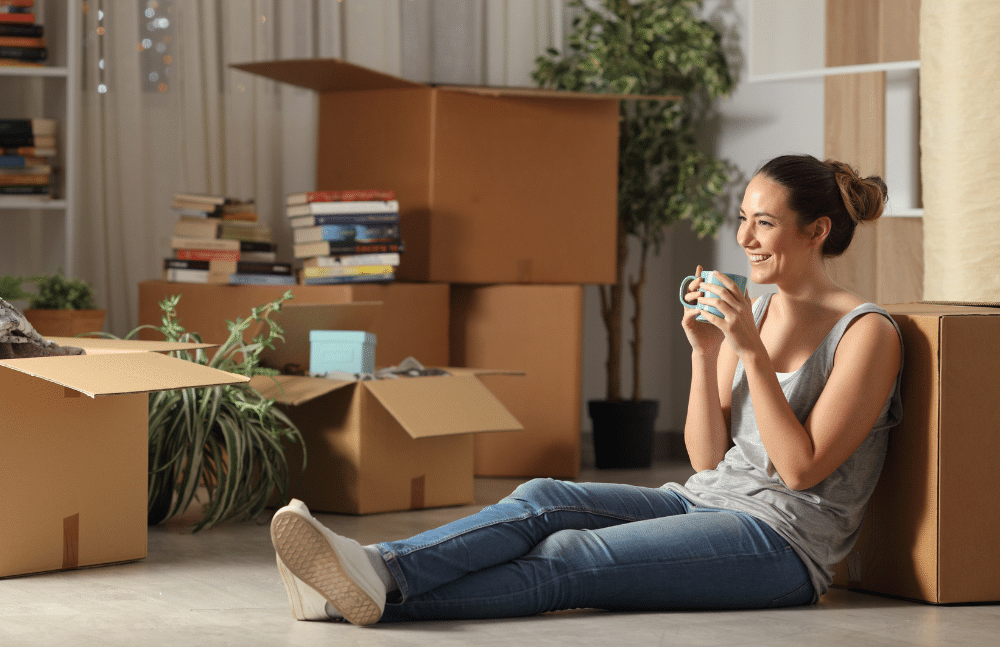 A woman sitting on the ground drinking coffee surrounded by boxes.