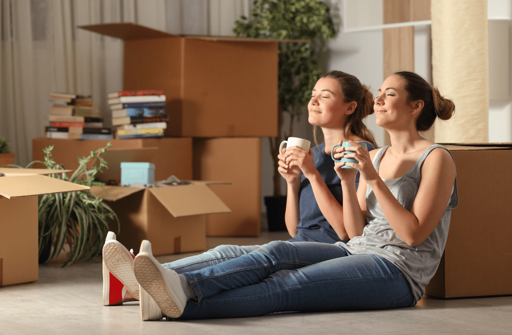 Two women sitting down drinking coffee surrounded by boxes.