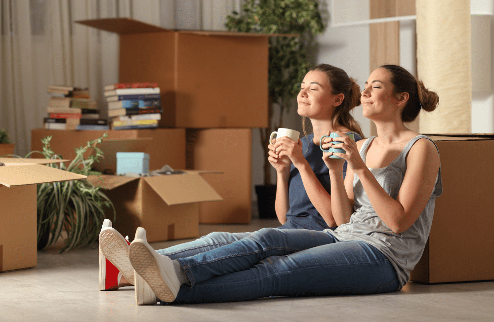 Two women sitting on the ground drinking coffee surrounded by boxes.