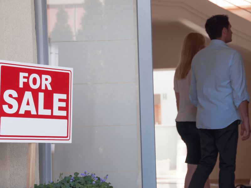 A man and a woman walking into a house with a for sale sign in the front.