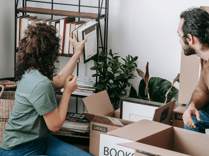 Couple unpacking books from a cardboard box and putting the books on a shelf.