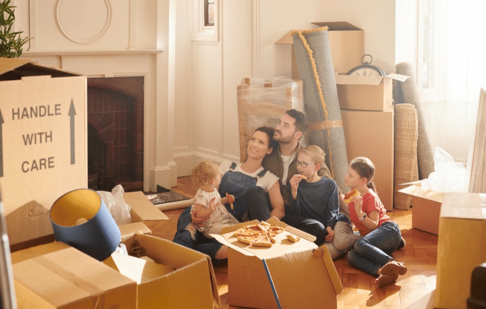 Family sitting on the floor eating pizza surrounded by cardboard moving boxes and rolled up rugs.