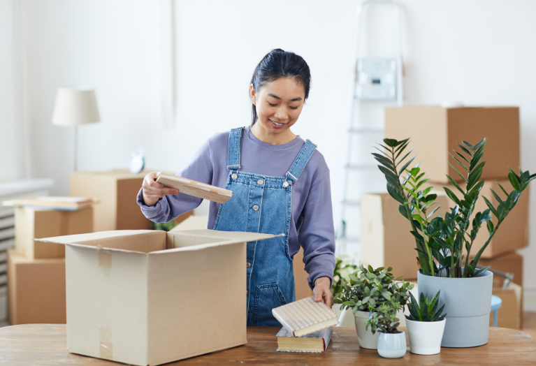Woman unpacking notebooks from a cardboard box and placing them next to potted plants on a table.