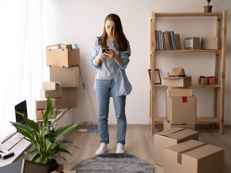 Woman standing in the middle of the room looking at her phone surrounded by cardboard boxes.