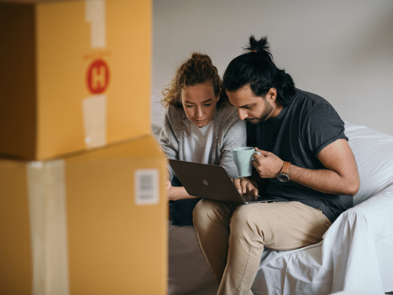 Man and women hunched over their laptop while sitting on a couch with cardboard boxes in front of them.