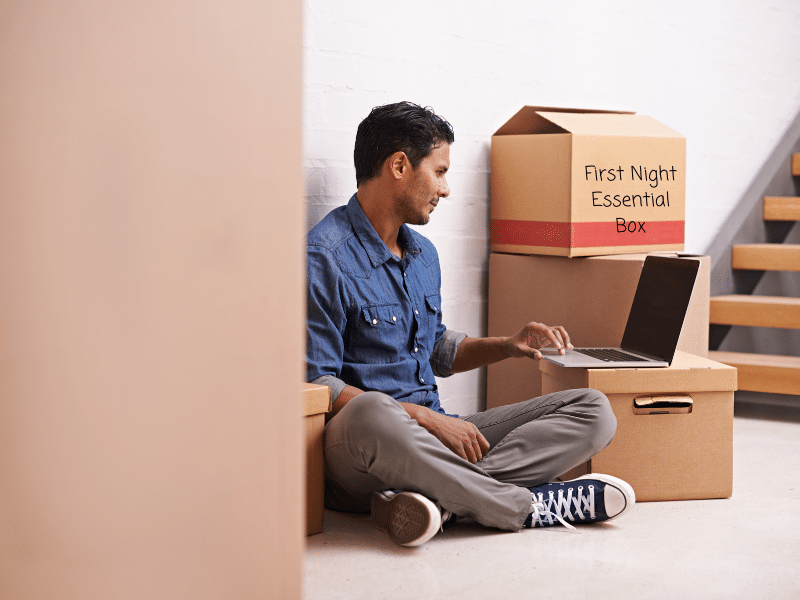 Man sitting on the floor next to stairs working on his laptop with a cardboard box labeled First Night Essential Box behind him.